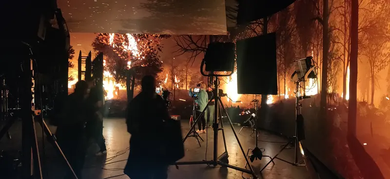 Crew and lighting stands in front of an LED volume showing a forest on fire, casting orange light across the set.